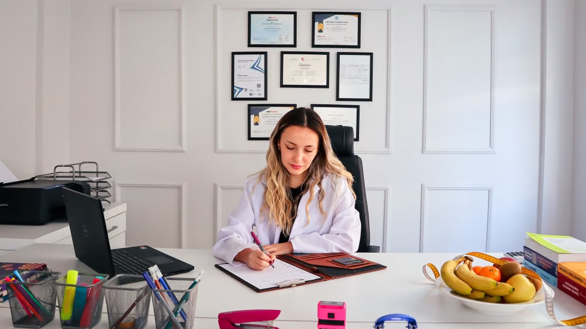 Nutritionist sits at a modern office desk writing, with fruits and diplomas displayed, emphasizing health and professionalism.