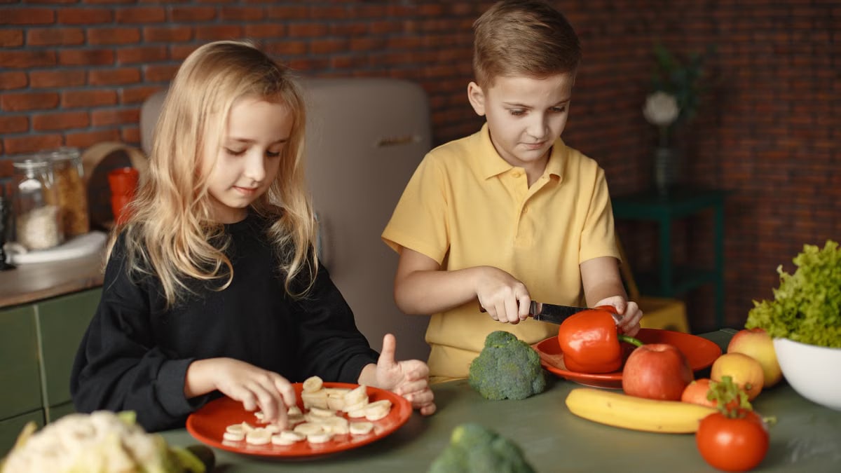 Focused little girl and boy cutting various vegetables and fruits while sitting at table and cooking in loft kitchen with brick wall
